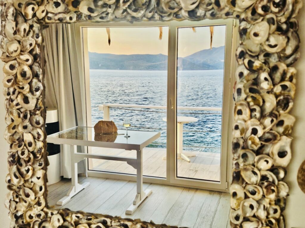 Reflection of a peaceful seaside view through a shell-framed mirror, with a table, wine glass, and serene waters beyond the glass doors